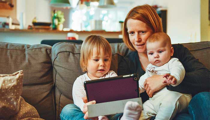 A mother and her children sit together on a couch in a cozy living room with a tablet